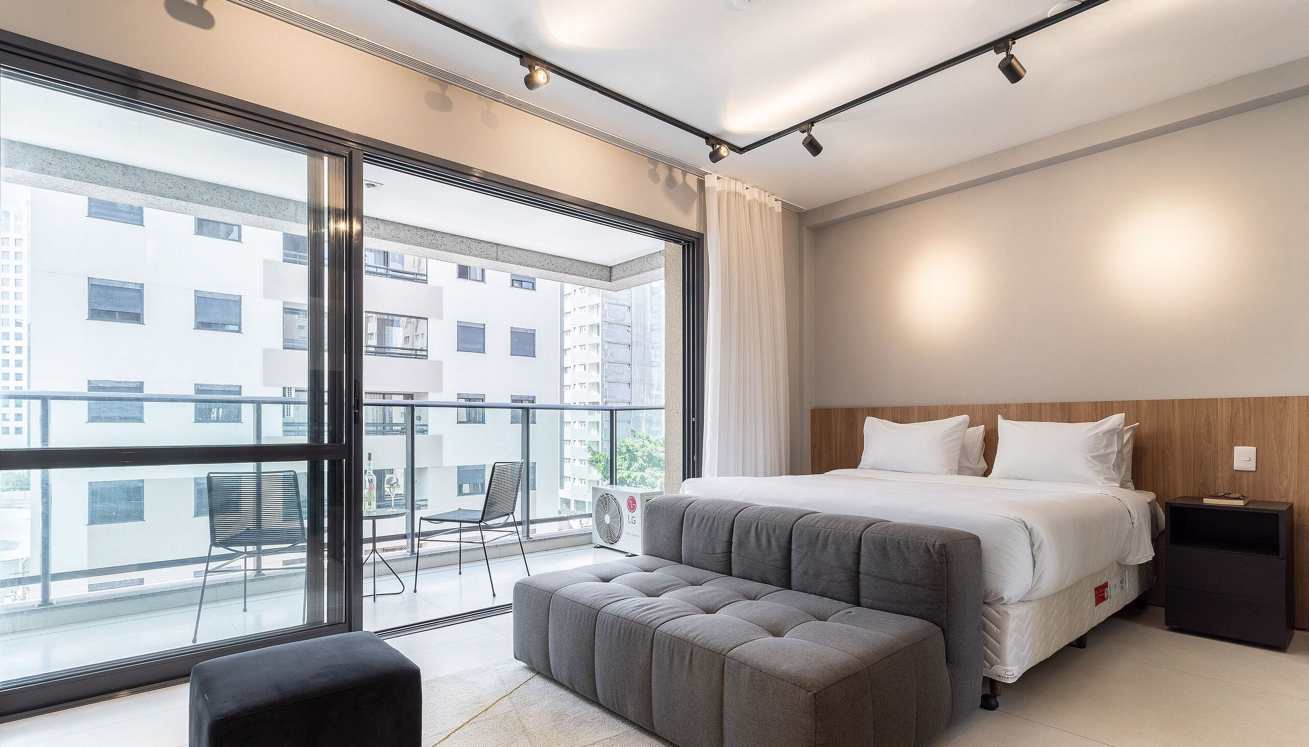A modern hotel room with a large balcony, a bed with white linens, a grey upholstered bench, and a small table with a fan.