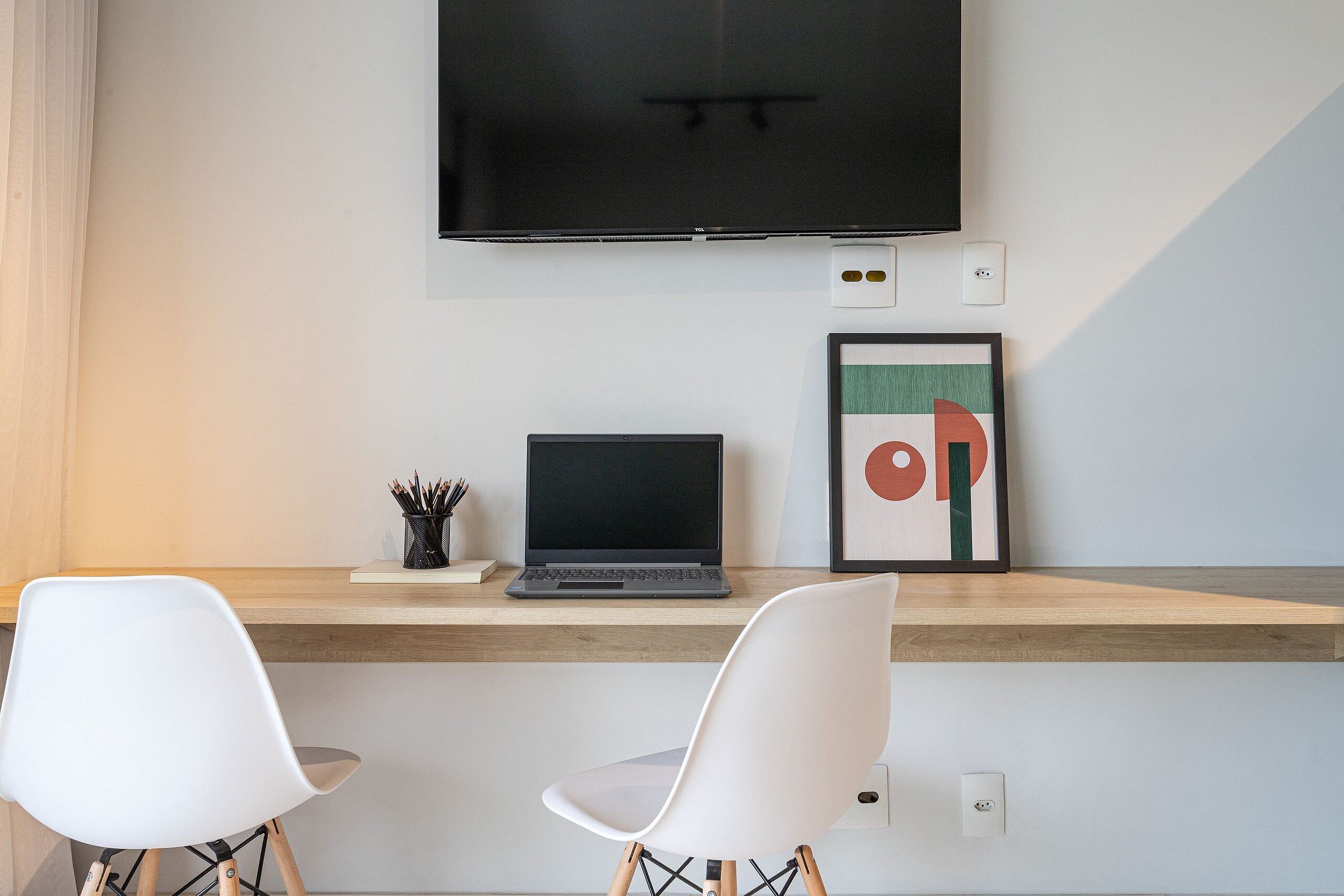 A modern workspace with a laptop on a wooden desk, two white chairs, a framed abstract art piece, and a mounted TV.