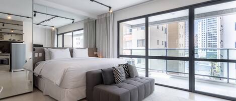 A modern bedroom with a large bed, a grey ottoman, and a view of the city through large windows.