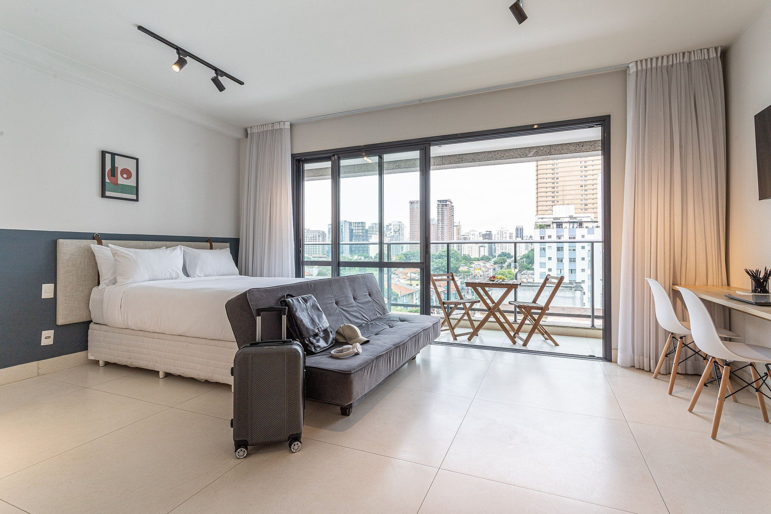 Ein Hotelzimmer mit einem Bett, einem grauen Sofa, einem Schreibtisch mit Stühlen und einem großen Fenster mit Blick auf die Stadt.