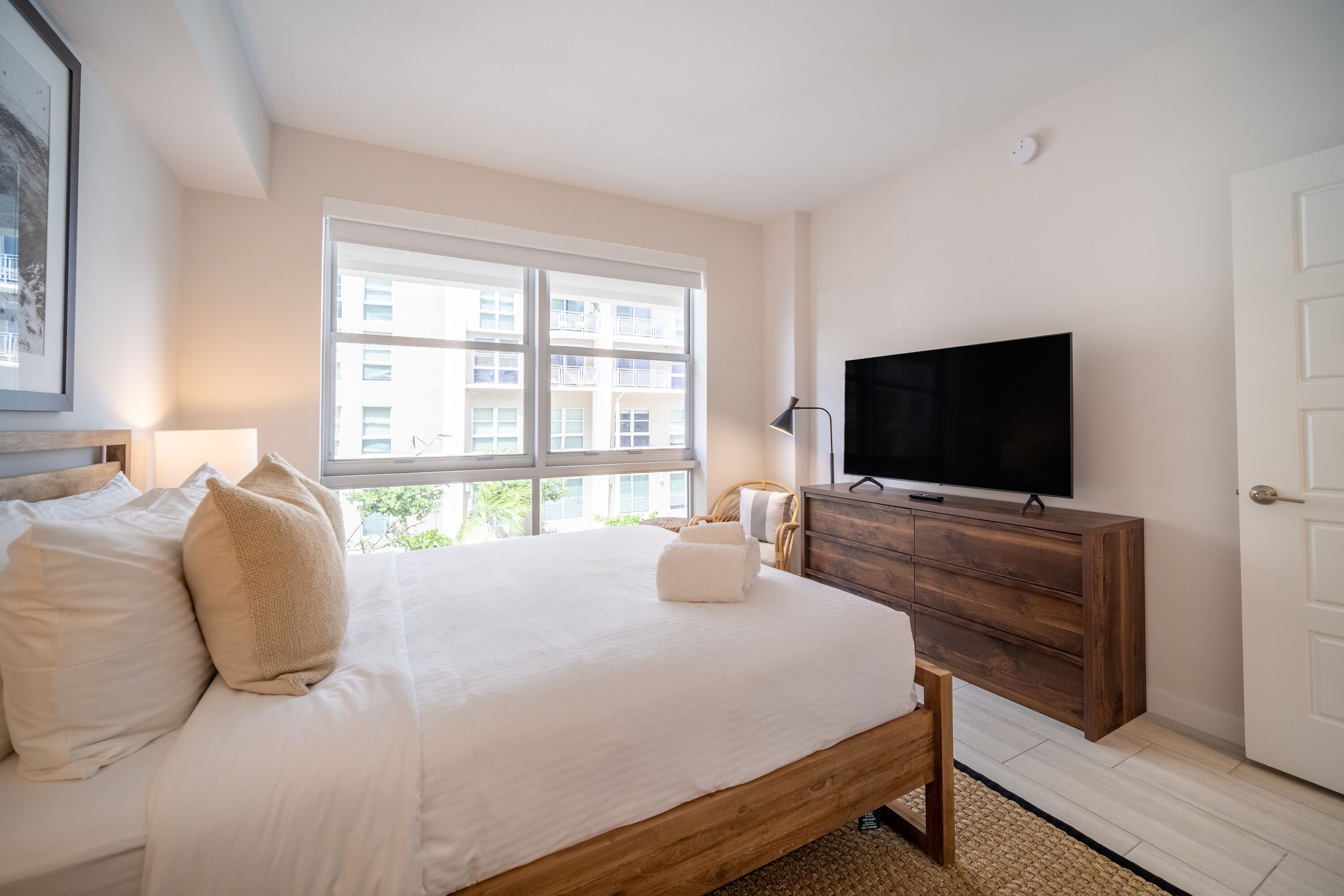 A bedroom with a large bed, a wooden dresser, a television, and a window with a view of buildings.