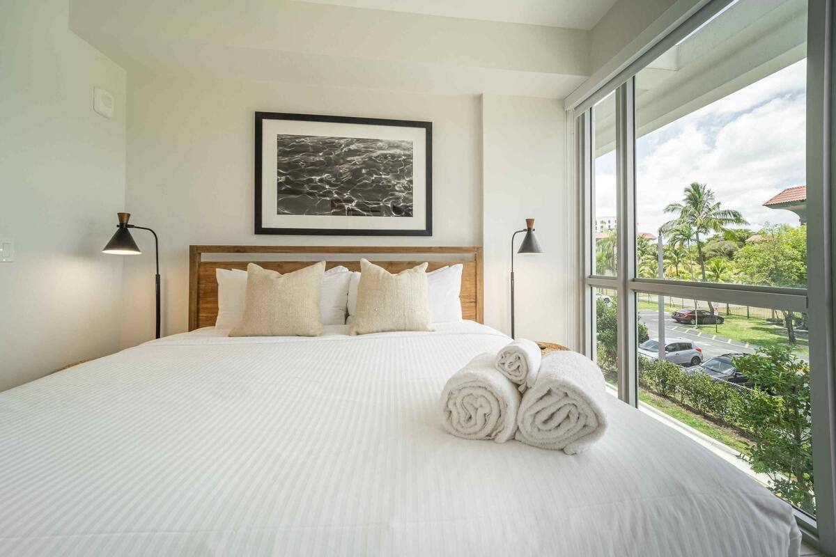 A neatly made bed with white linens and rolled towels, a framed picture on the wall, and a view of a parking area through a large window.