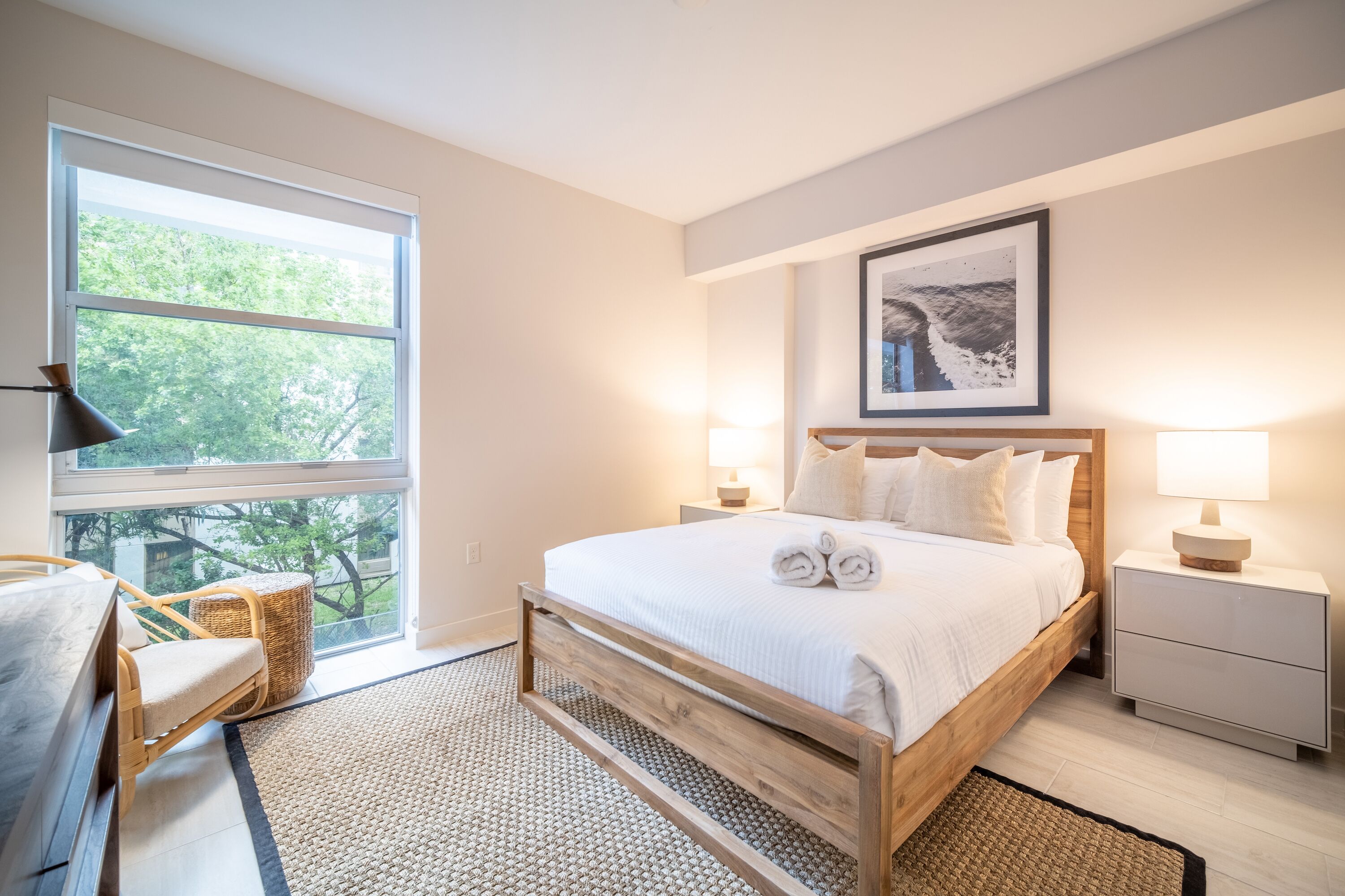 A bedroom with a wooden bed, two bedside lamps, a large window, a wicker chair, and a framed picture on the wall.