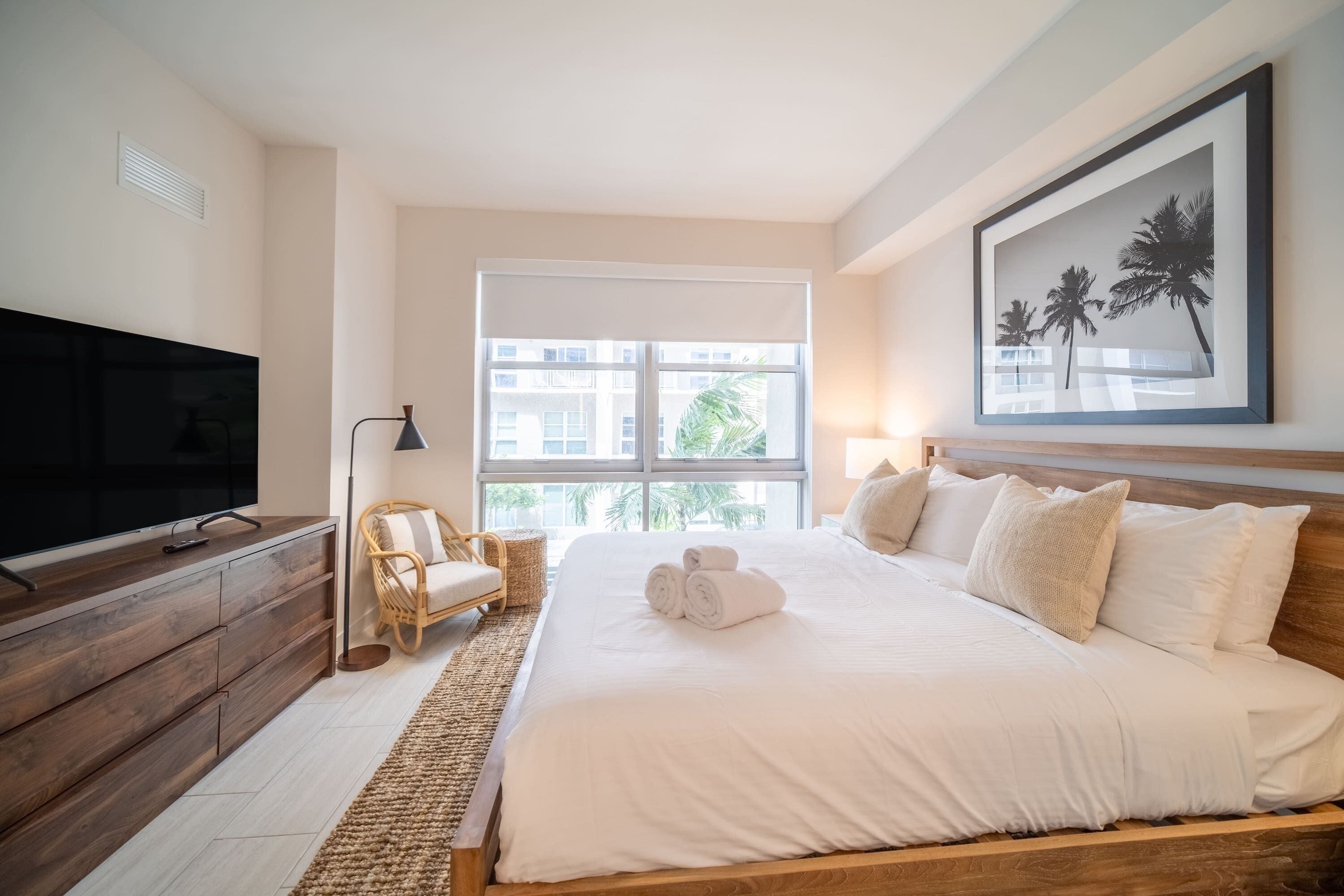 Ein Schlafzimmer mit einem großen Bett, einem Fernseher, einem Korbstuhl und einem Fenster mit Blick auf Gebäude und Palmen.