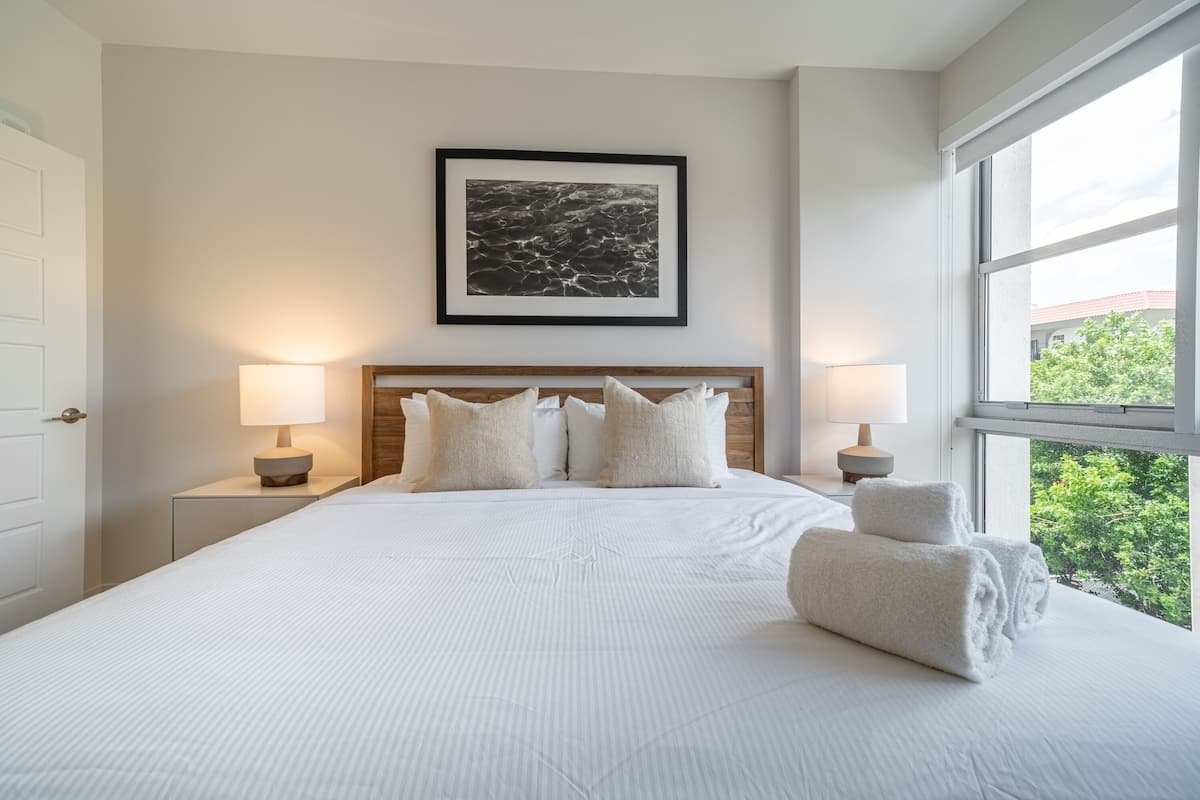A neatly made bed with white linens and pillows, a framed artwork above it, and a view of greenery through the window.