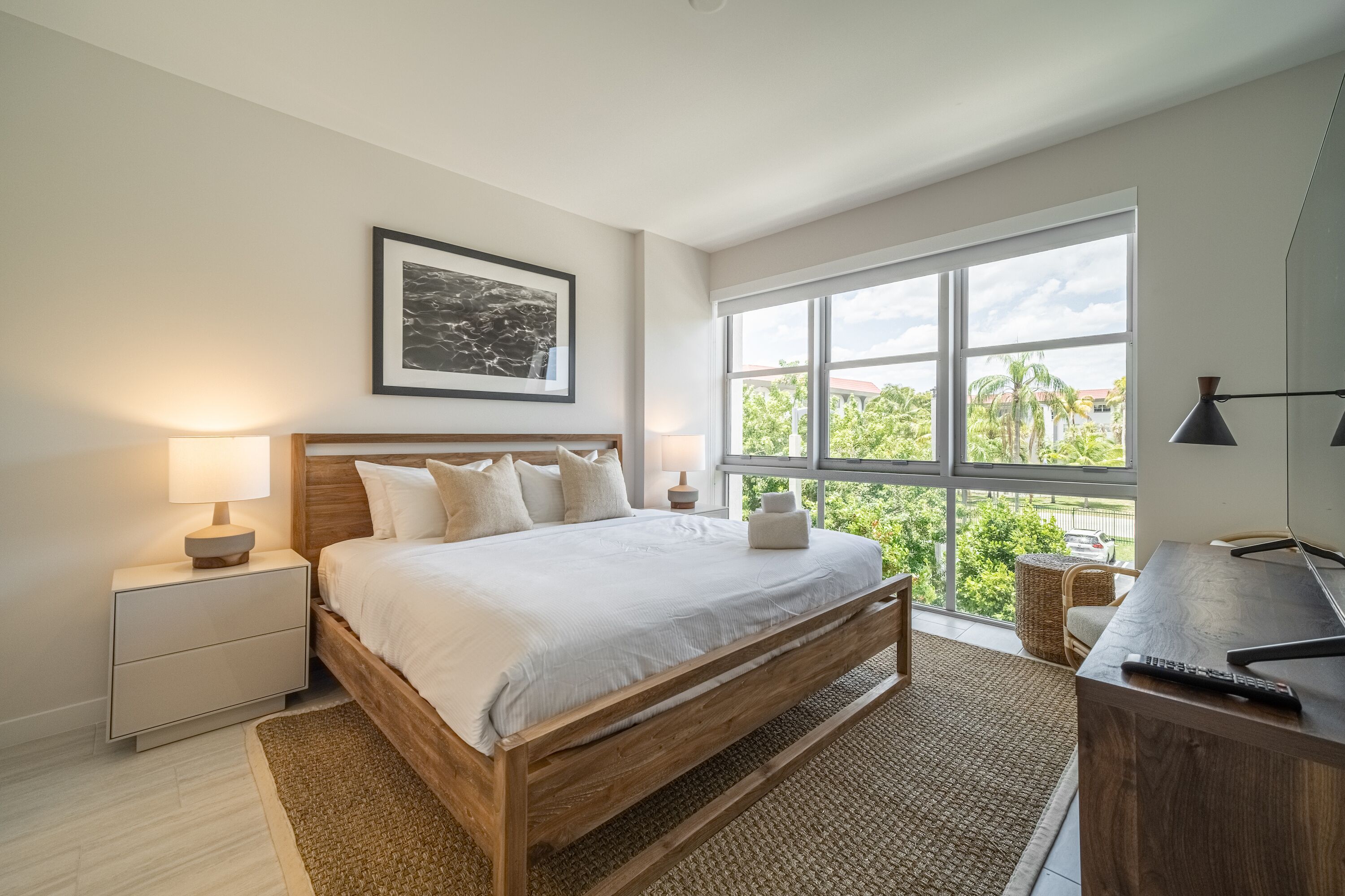 A bedroom with a wooden bed, white bedding, a nightstand with a lamp, a desk with a computer, and a large window with a view of greenery.