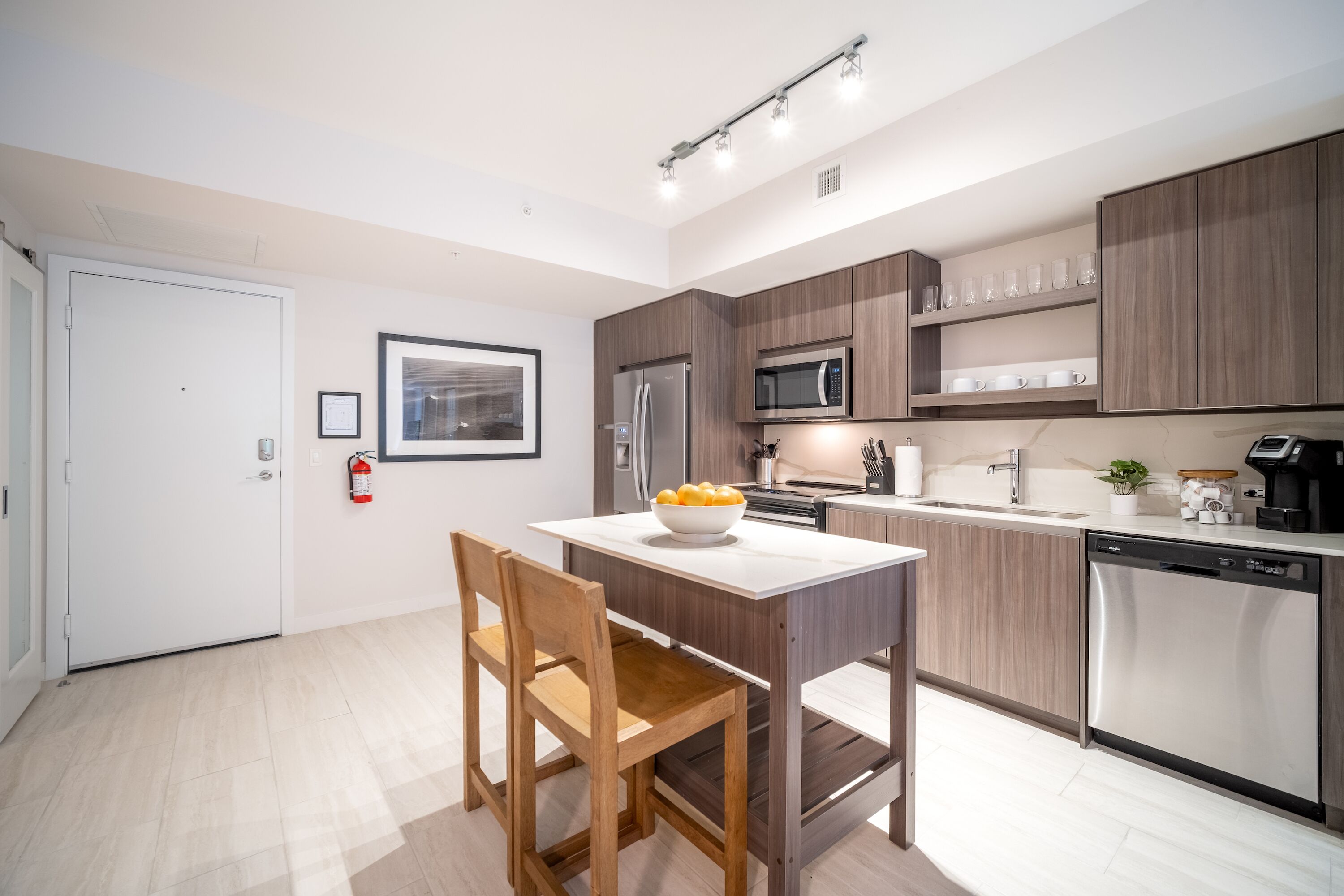 A modern kitchen with wooden cabinets, a stainless steel refrigerator, a microwave, and a dishwasher. There is a dining table with a bowl of fruit and a chair.
