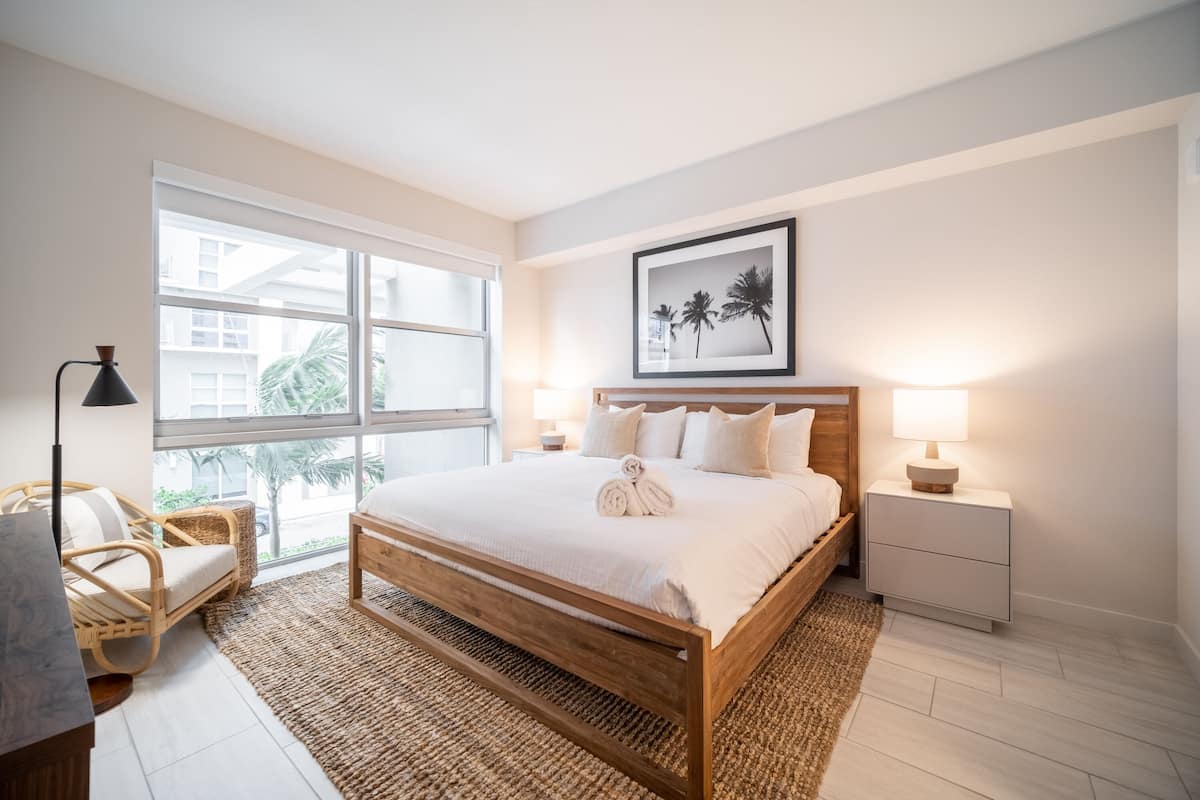 A bedroom with a wooden bed, a large window, a framed picture, and a nightstand with a lamp.