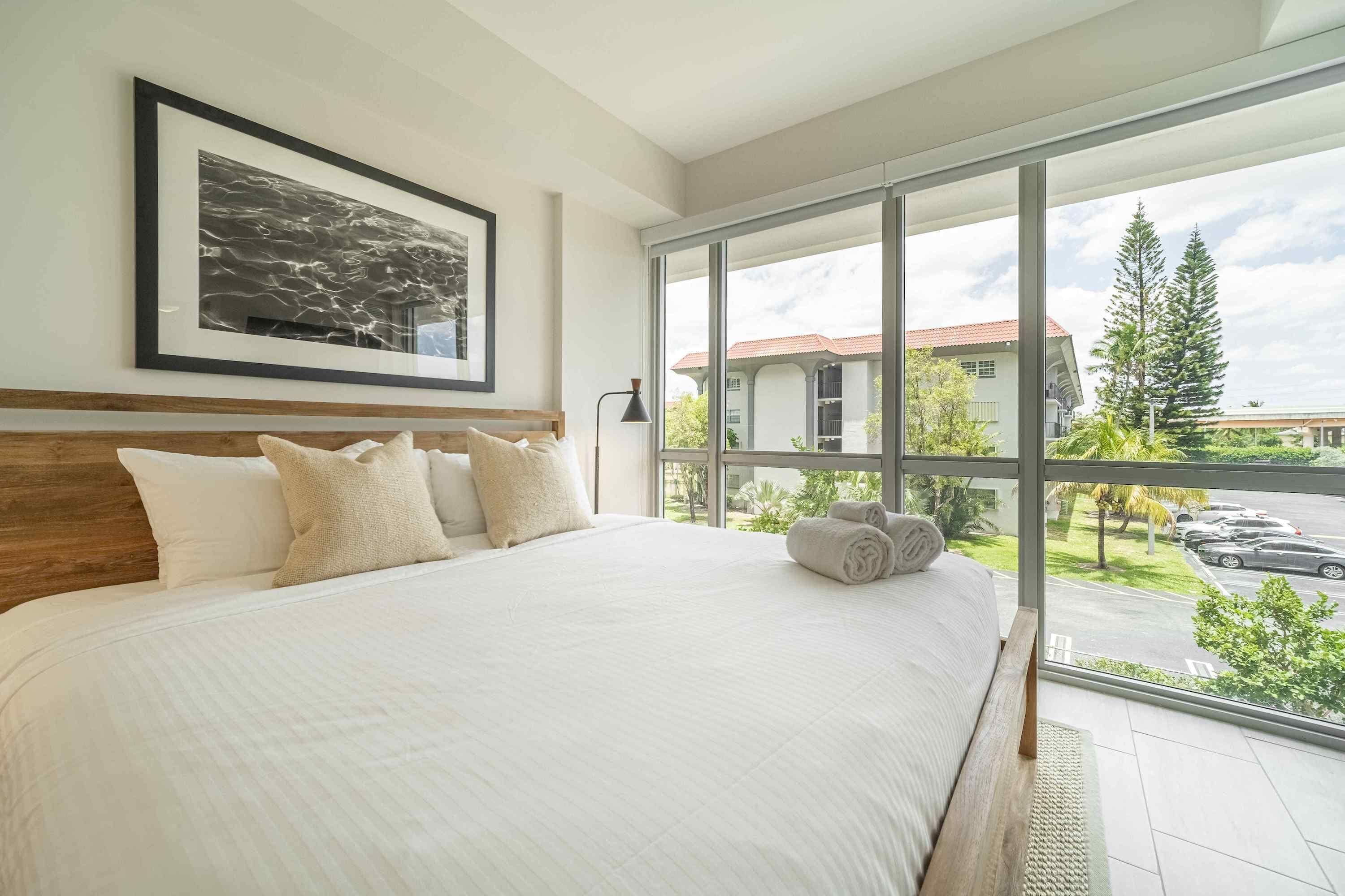 A bedroom with a large bed, a framed artwork above it, and a view of a street and trees through a large window.