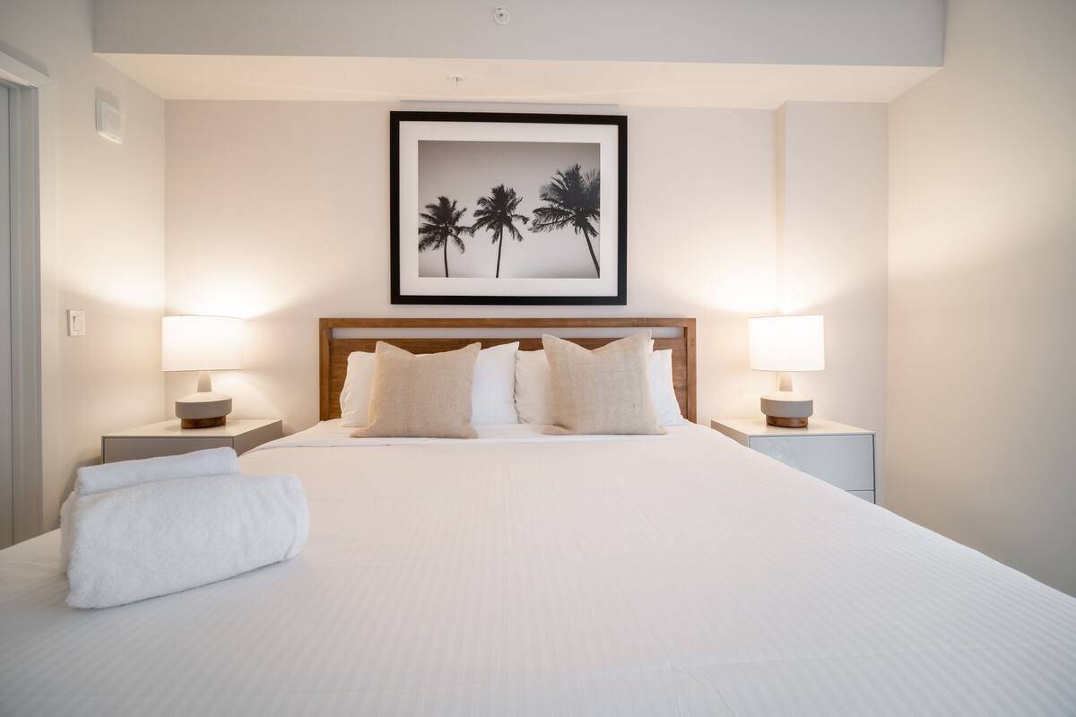 A neatly made bed with white linens and a rolled white towel, flanked by two bedside tables with lamps, in a room with a framed picture of palm trees on the wall.