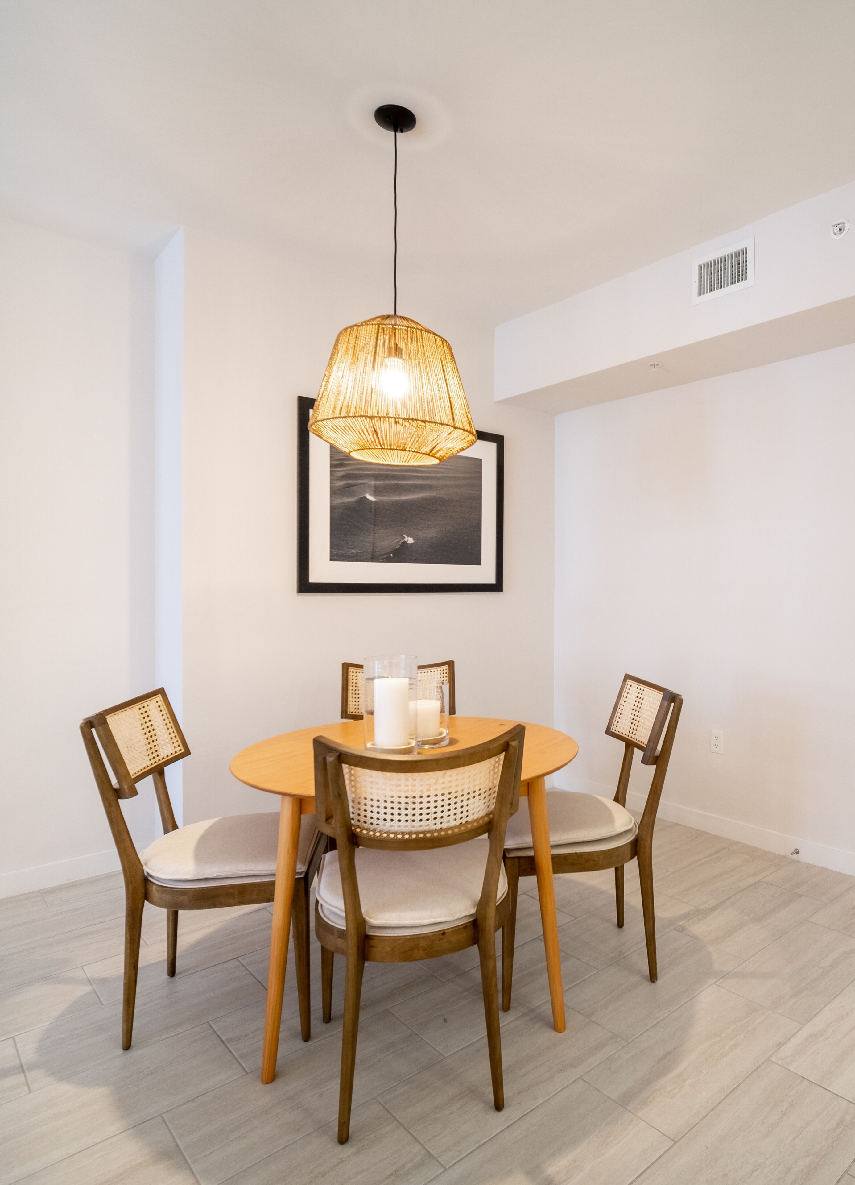 A dining area with a round table, four wooden chairs with woven seats, a hanging wicker pendant light, and a framed picture on the wall.