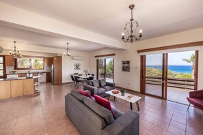 A spacious living area with a grey sofa, a dining table, and a kitchen in the background. - Villa Korona - Three Bedroom Villa, Sleeps 6 (Neo Chorio)
