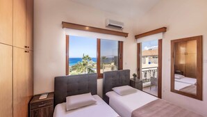 A bedroom with two beds, a wooden wardrobe, a nightstand, and a large window offering a view of the sea and greenery. - Villa Korona - Three Bedroom Villa, Sleeps 6 (Neo Chorio)