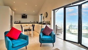 A modern living room with two blue armchairs, a red and a purple pillow, a dining area, and a large sliding glass door leading to a balcony with a city view. - Anchor View (Teignmouth)