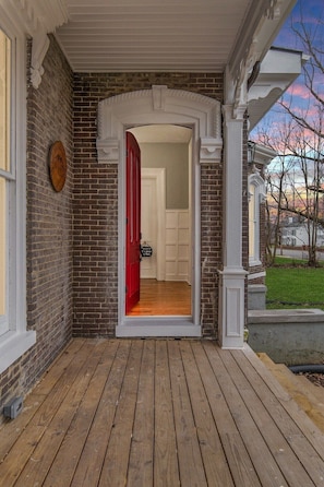 A wooden deck leading to a red door, with a brick wall and a white door visible in the background.
