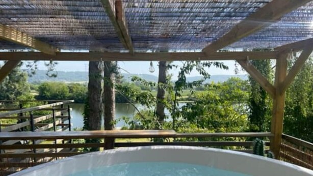 A hot tub with a wooden deck and a pergola overhead, overlooking a lake and greenery.
