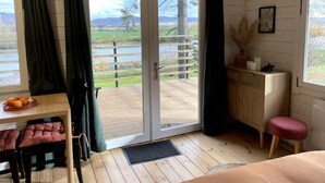 A room with a wooden floor, a desk with a basket of oranges, a bench, a small stool, a cabinet, and a view of a lake through a sliding door.