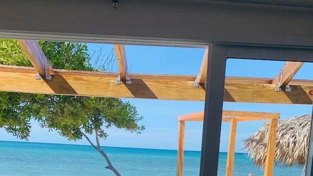 Uitzicht op een strand met een houten terras, een constructie met rieten dak en een bed met een gestreept kleed.