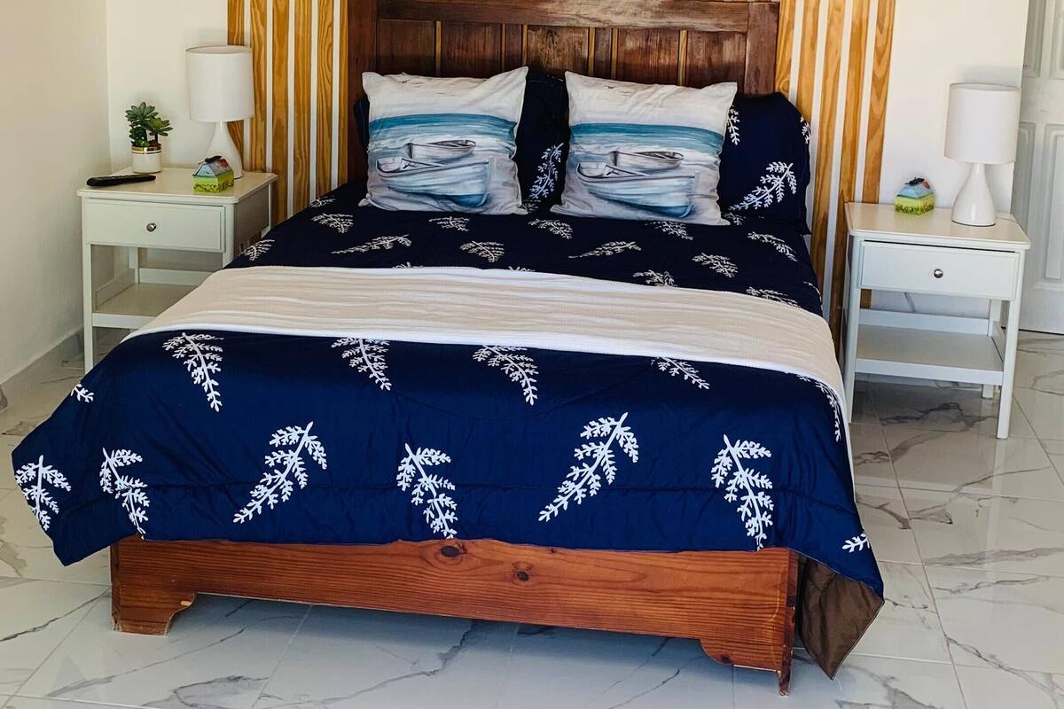 A bedroom with a wooden headboard, a bed with a blue bedspread, two bedside tables with lamps, and a ceiling fan.