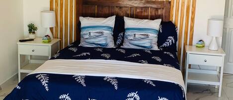 Un dormitorio con cabecera de madera, cama con cubrecama azul, dos mesitas de noche con lámparas y un ventilador de techo.