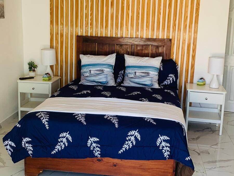 Quarto com cabeceira de madeira, cama com colcha azul, duas mesas de cabeceira com abajures e um ventilador de teto.