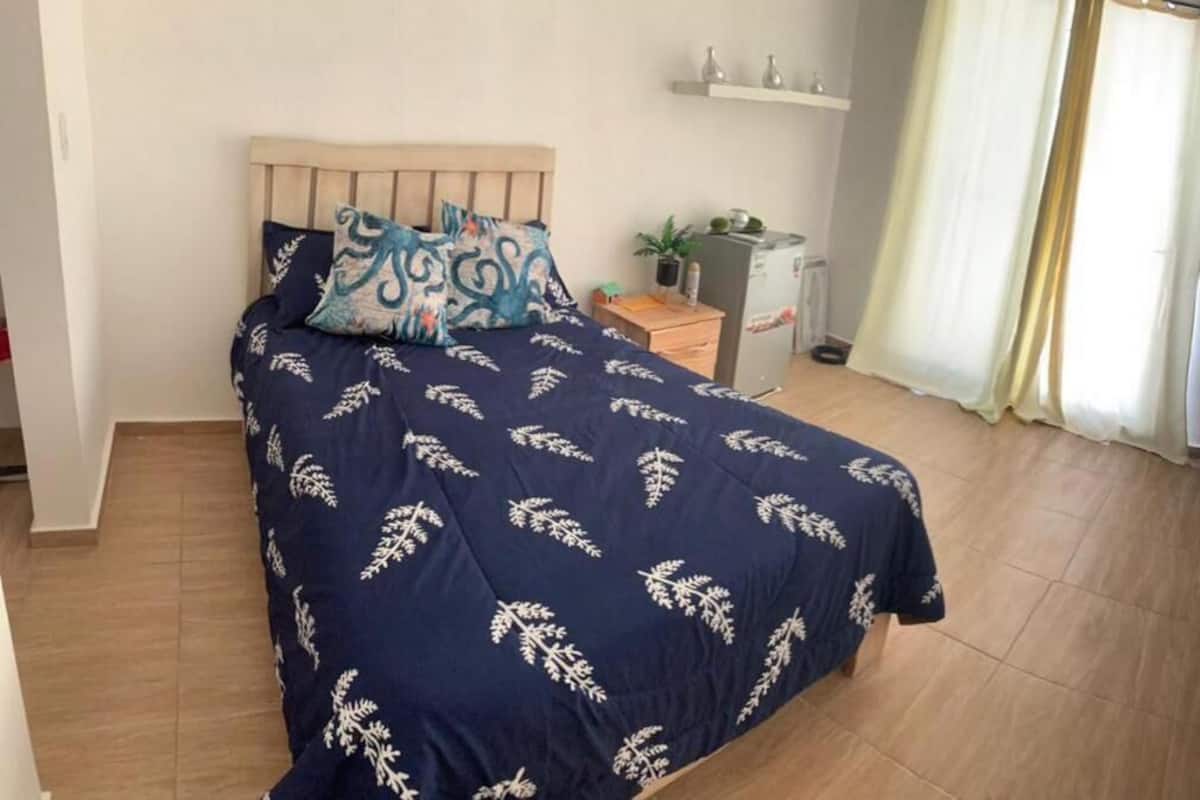 A bedroom with a bed, a nightstand, a small table with a plant, a desk, and a wardrobe.