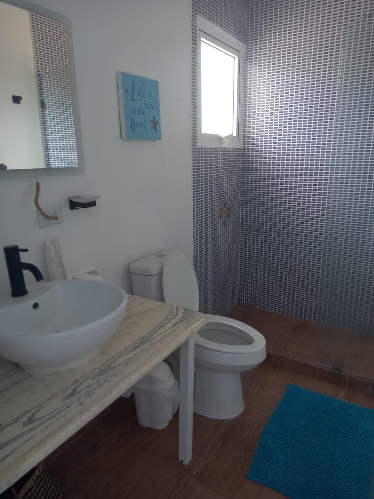 A bathroom with a white sink, toilet, and shower. There is a blue sign on the wall and a window above the shower.
