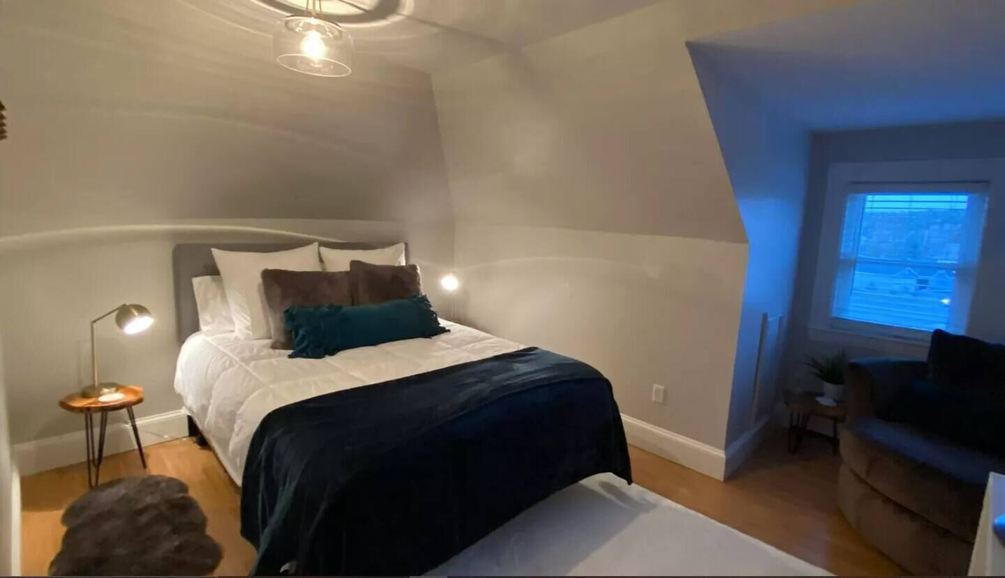 A bedroom with a bed, bedside table, lamp, and a window with blinds.