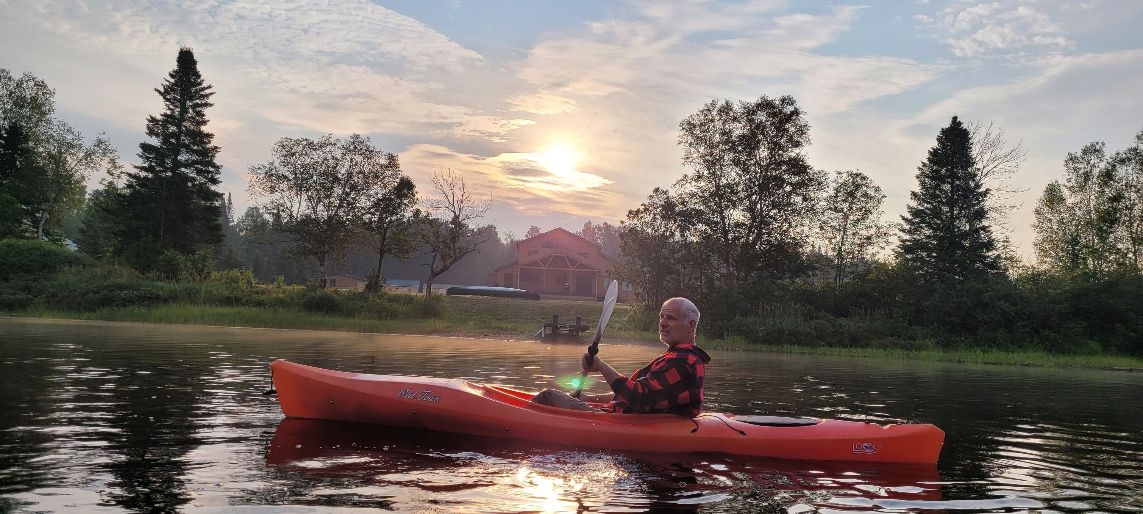 A person kayaking on a calm river with trees and houses in the background.