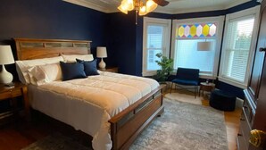 Un dormitorio con una cama grande, cabecera de madera, dos lámparas de noche, una mesita con lámpara, una silla y una ventana con persianas y un diseño de vitral.
