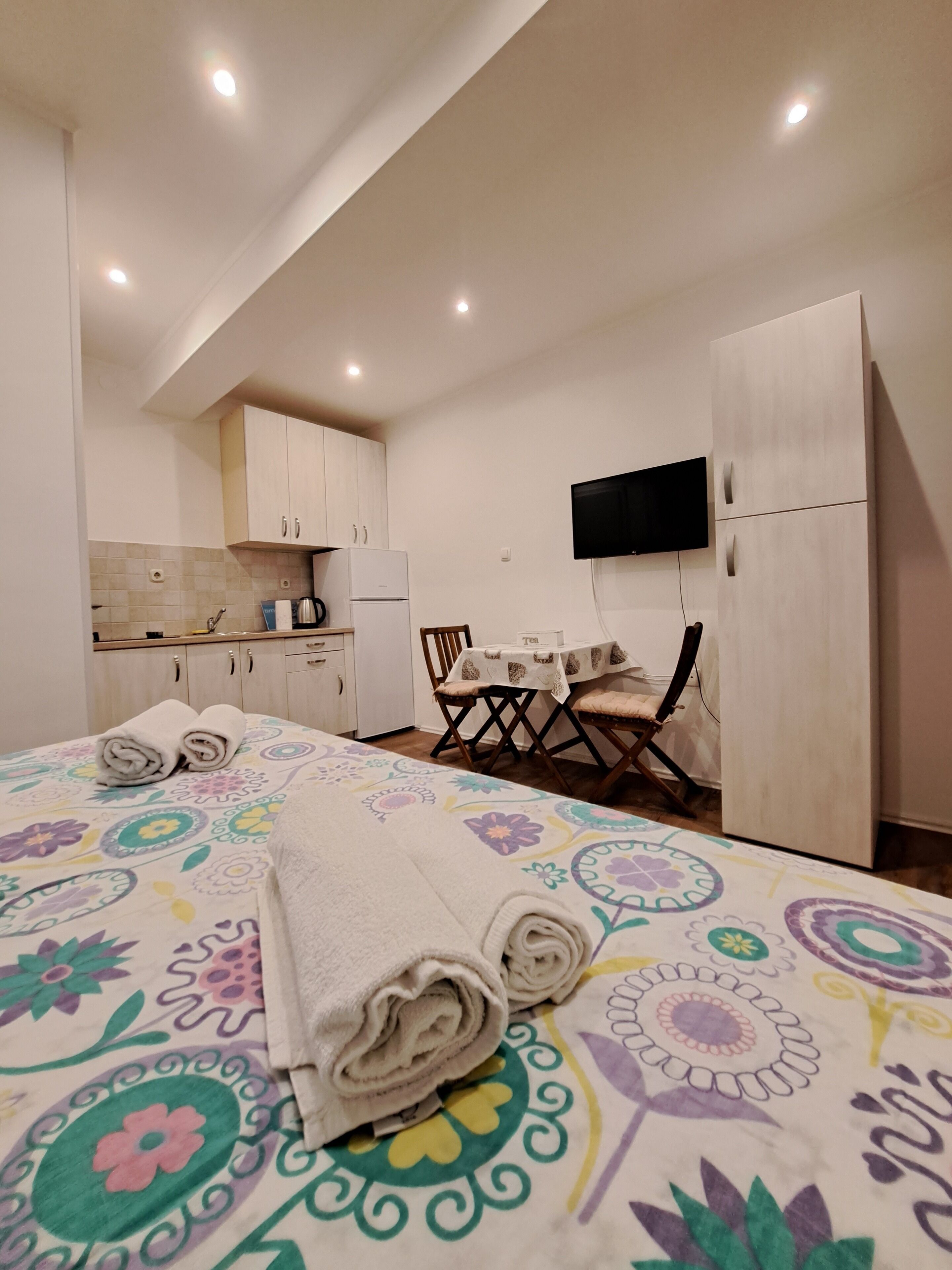 A compact hotel room with a bed, a small kitchenette, a dining area, and a TV mounted on the wall.