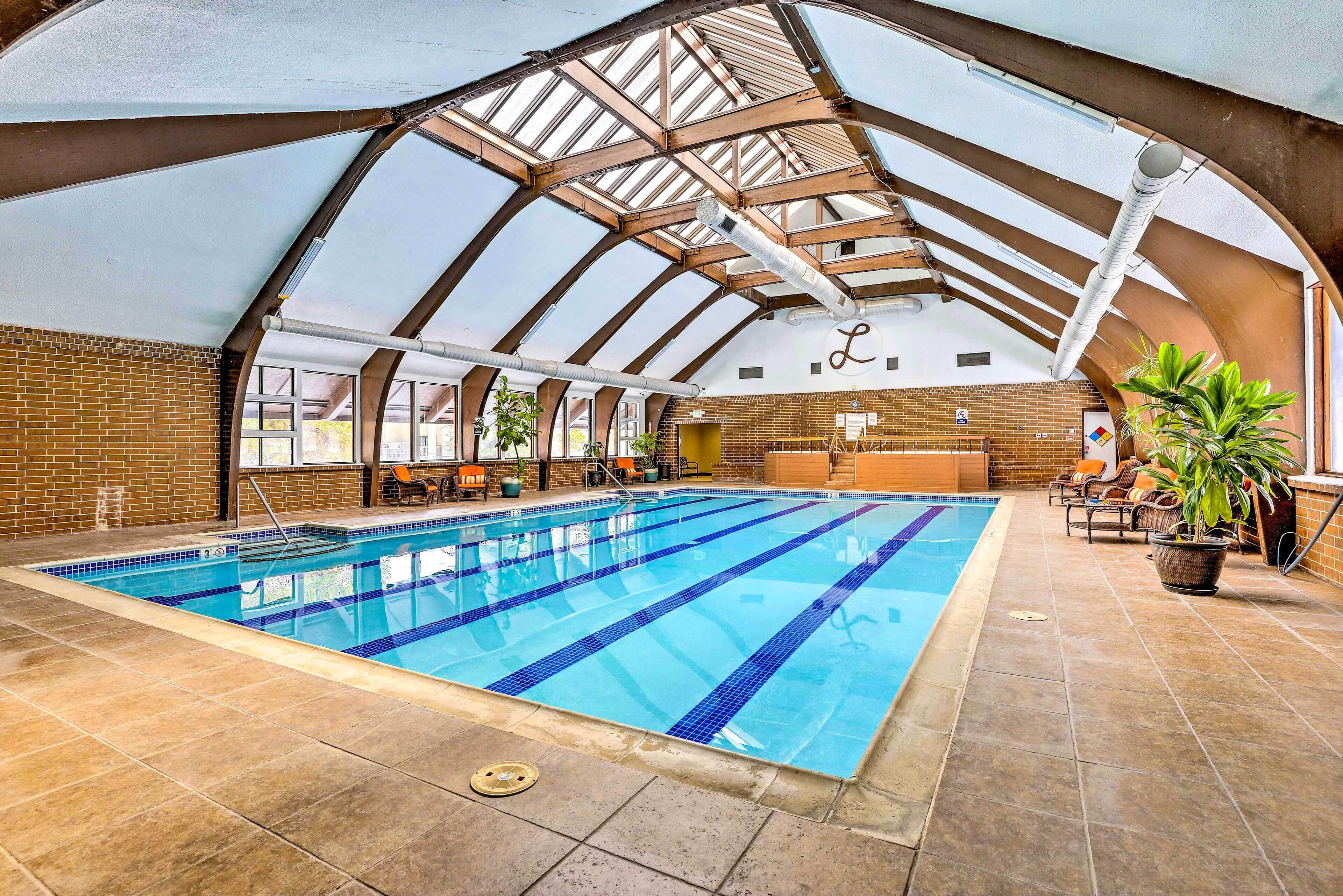 An indoor swimming pool with a high arched ceiling, tiled floor, and seating area with plants.