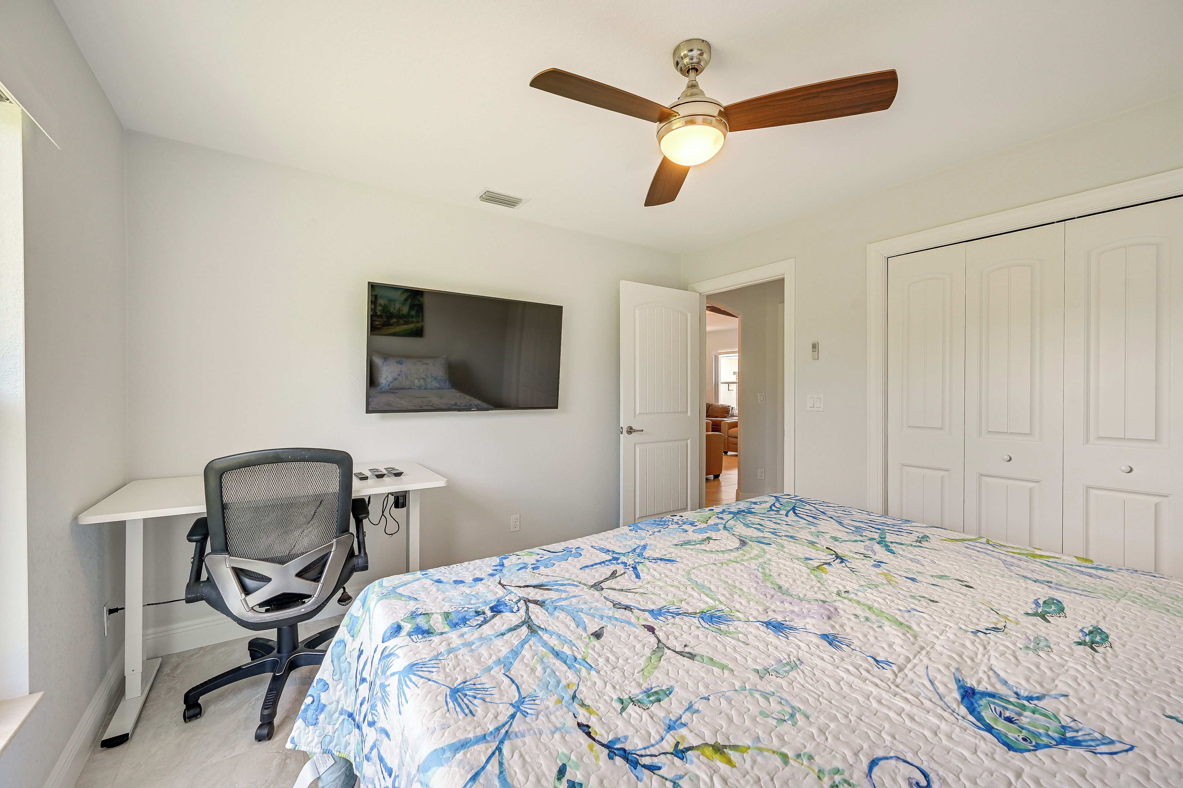 A bedroom with a bed, a desk, a chair, a ceiling fan, and a door leading to another room.