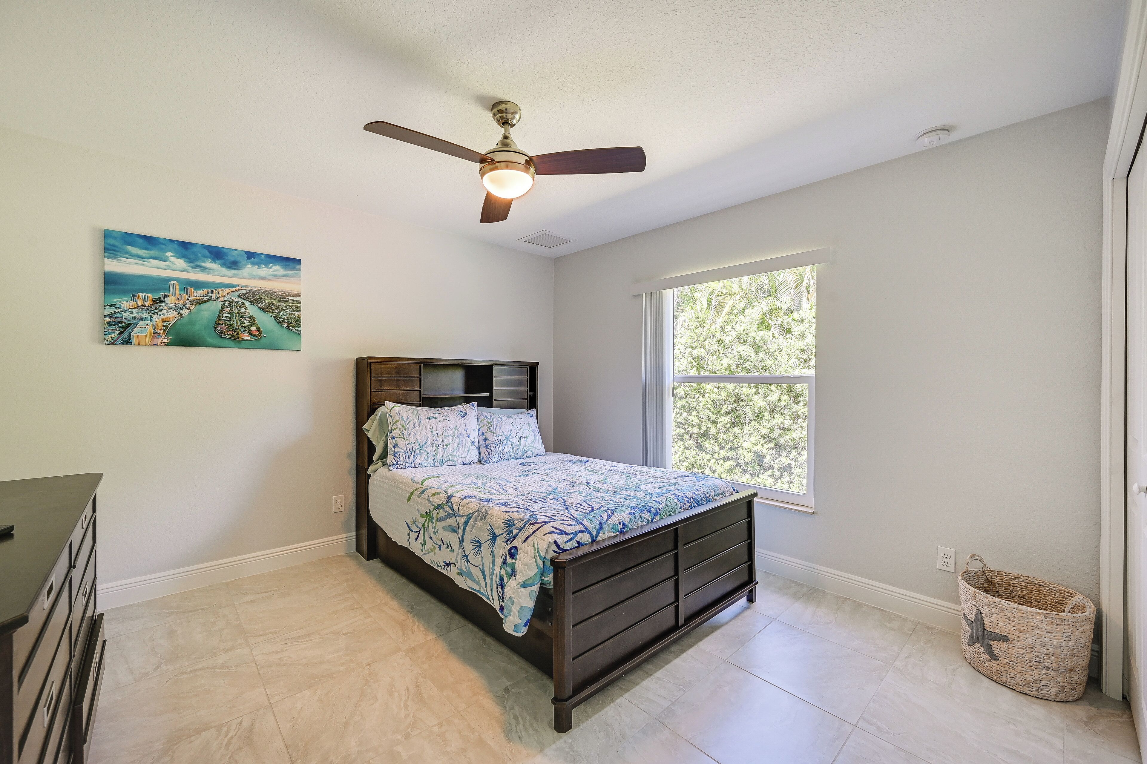 A bedroom with a bed, a ceiling fan, a window, a painting, and a small basket.