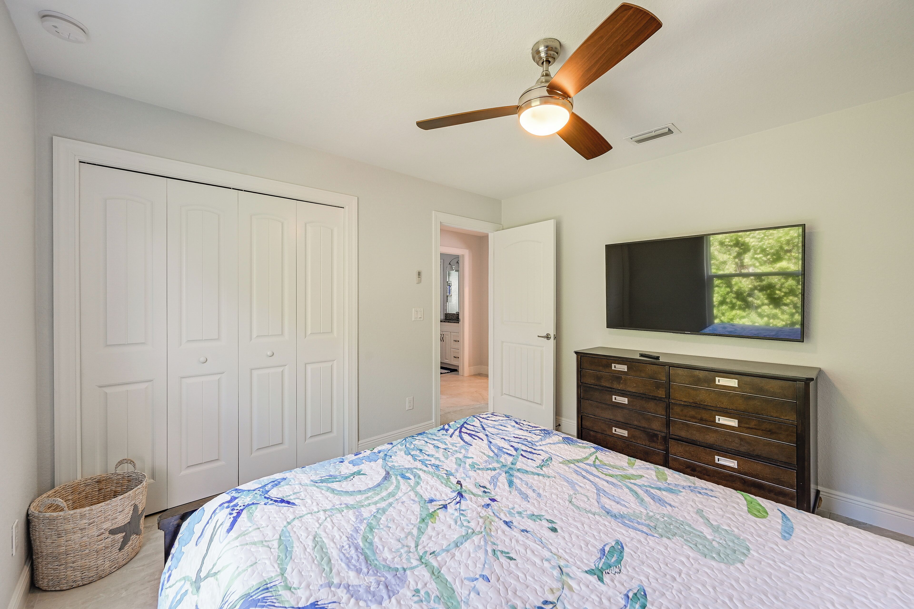A bedroom with a bed, a ceiling fan, a dresser, a TV, and a door leading to another room.