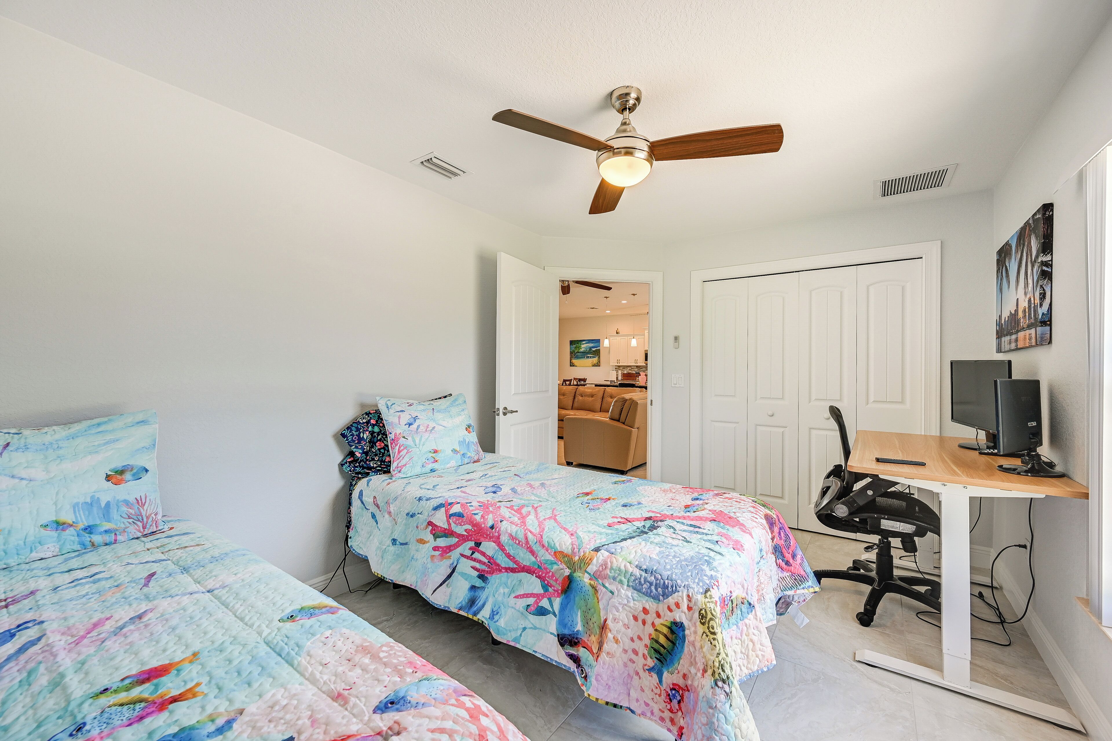 A bedroom with two beds, a desk with a computer, a ceiling fan, and a door leading to another room.