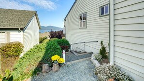 A house with a gravel driveway, a small garden with various plants, and a view of mountains in the background.
