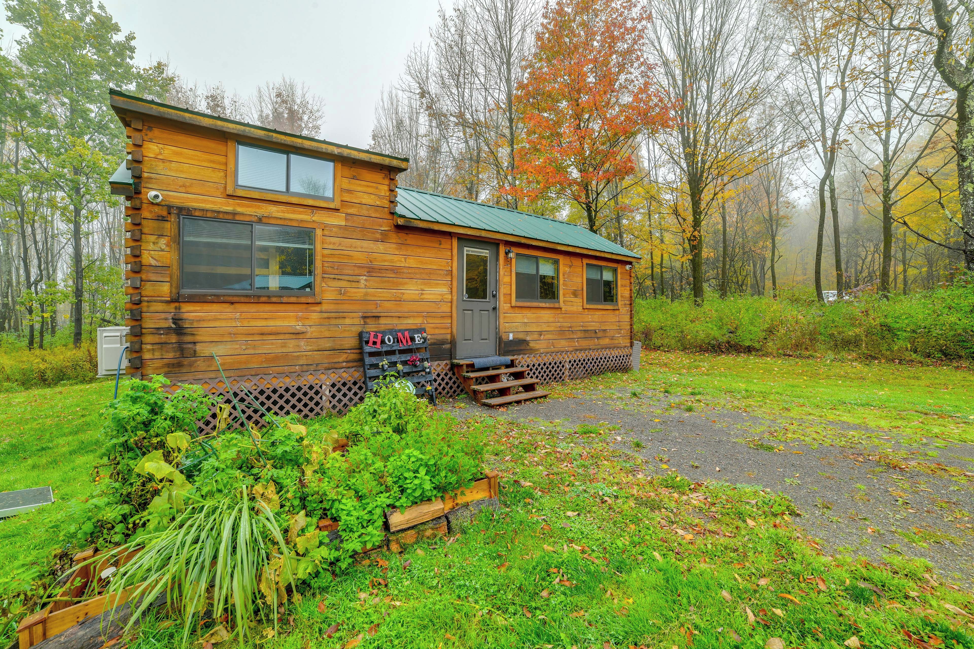Une cabane en bois avec un porche à l’avant, une petite terrasse et un jardin agrémenté de plantes et d’une clôture en bois.