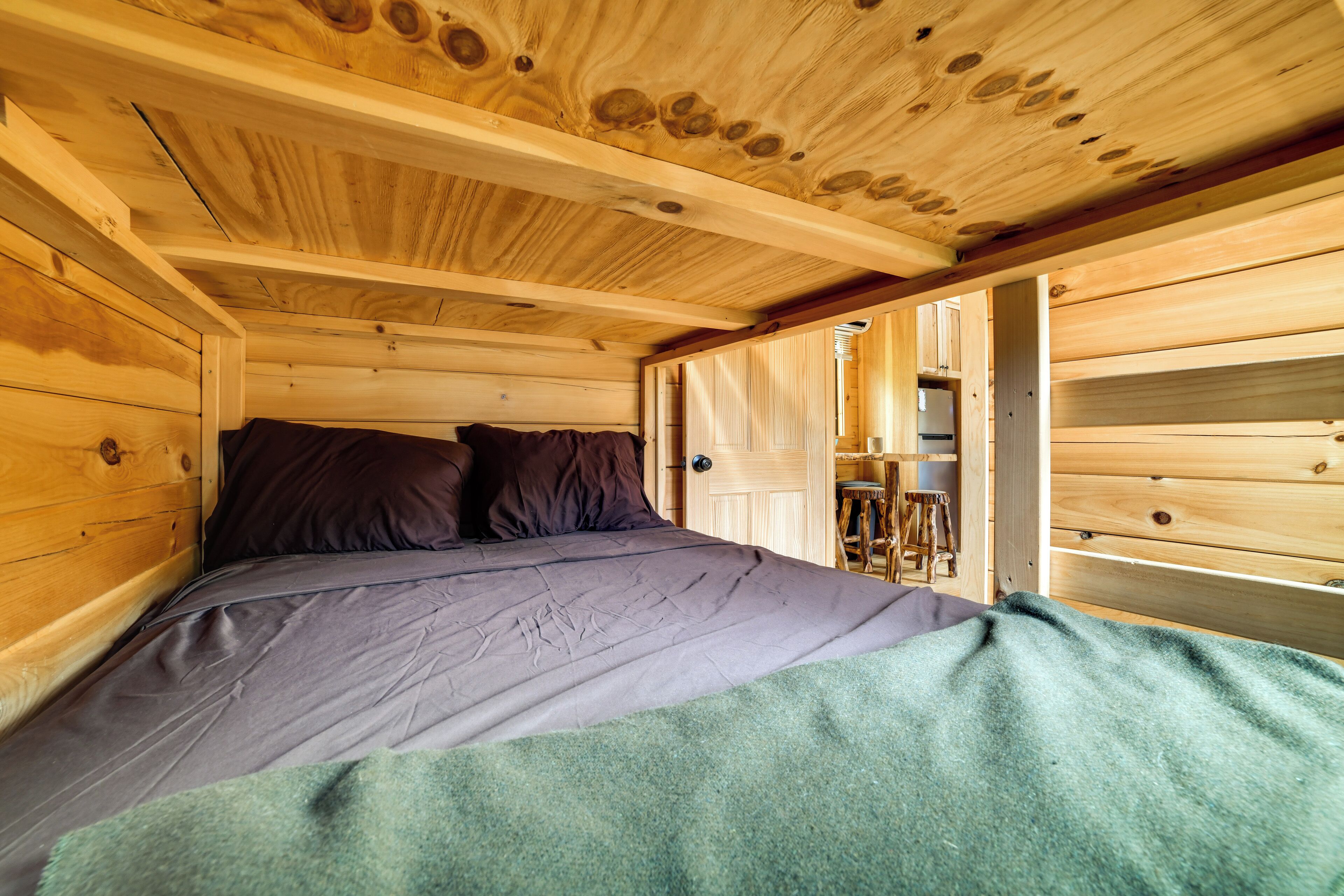 A wooden cabin interior with a bed, pillows, and a door leading to a kitchen area.