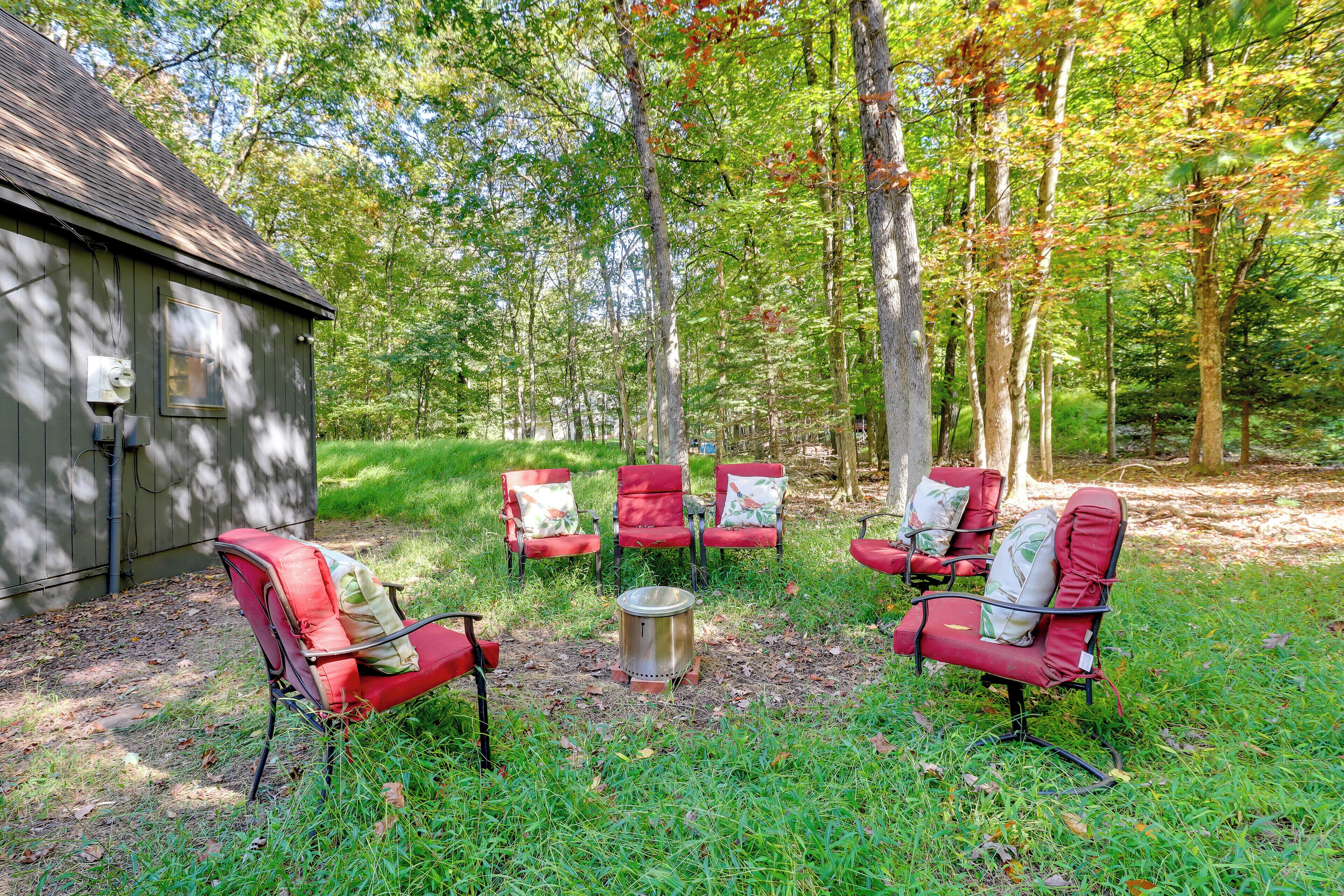 A rustic outdoor seating area with red chairs and a metal stove, surrounded by trees and a wooden building.