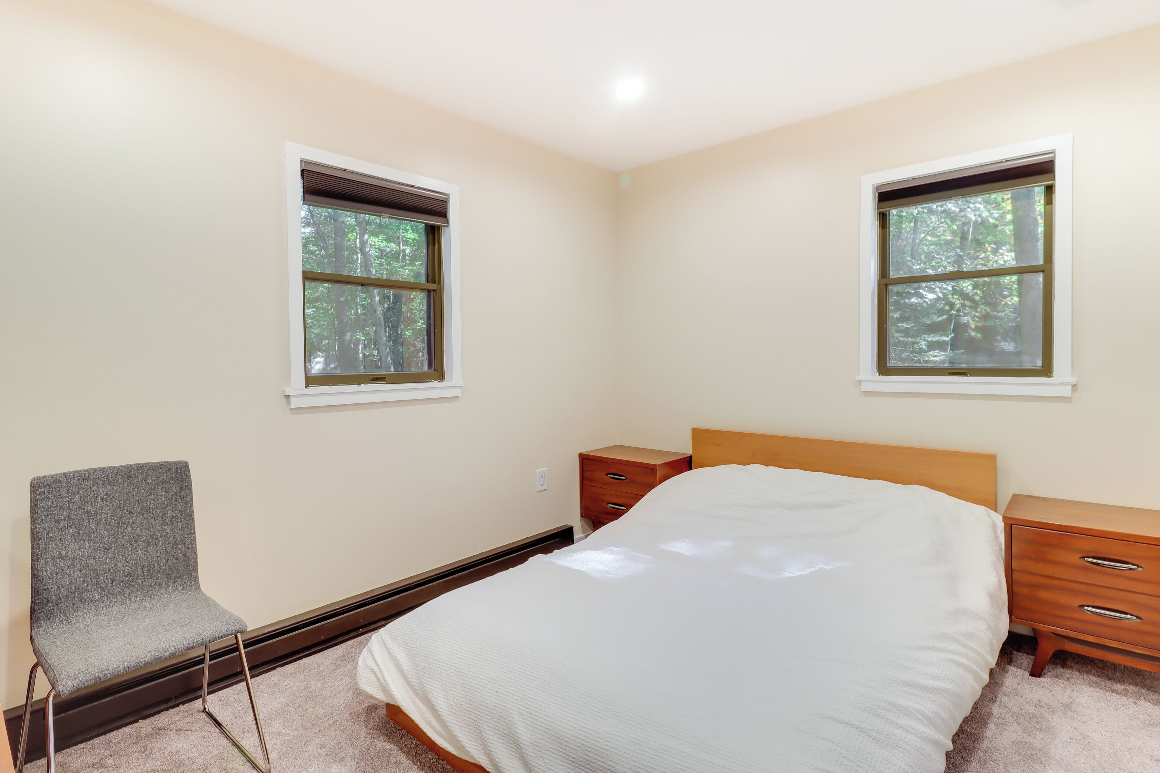 Quarto com cama, uma cadeira, duas mesas de cabeceira e duas janelas.