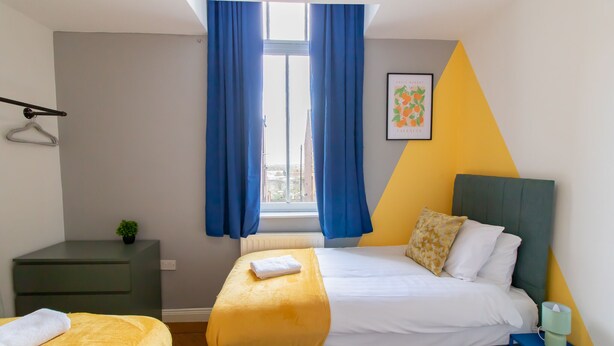 Un dormitorio con una cama, una mesita de noche, un pequeño mueble auxiliar, una ventana con cortinas y una obra de arte en la pared.