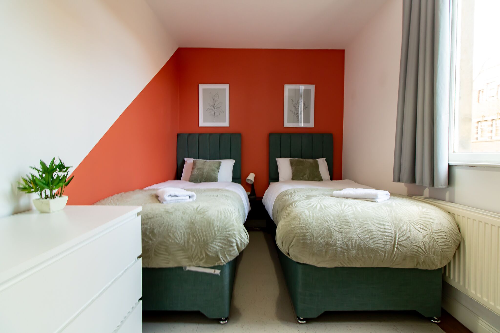 Une chambre avec deux lits, une commode blanche, une plante en pot et deux tableaux encadrés accrochés au mur.