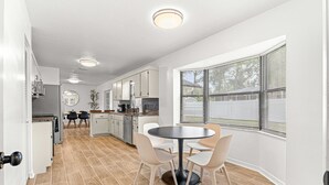 Une cuisine moderne avec des meubles blancs, un coin repas doté d’une table ronde et de chaises, et une grande fenêtre avec des stores.