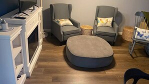 Een woonkamer met een televisie, twee fauteuils, een rond hocker, een open haard en een eethoek met een stoel en tafel.