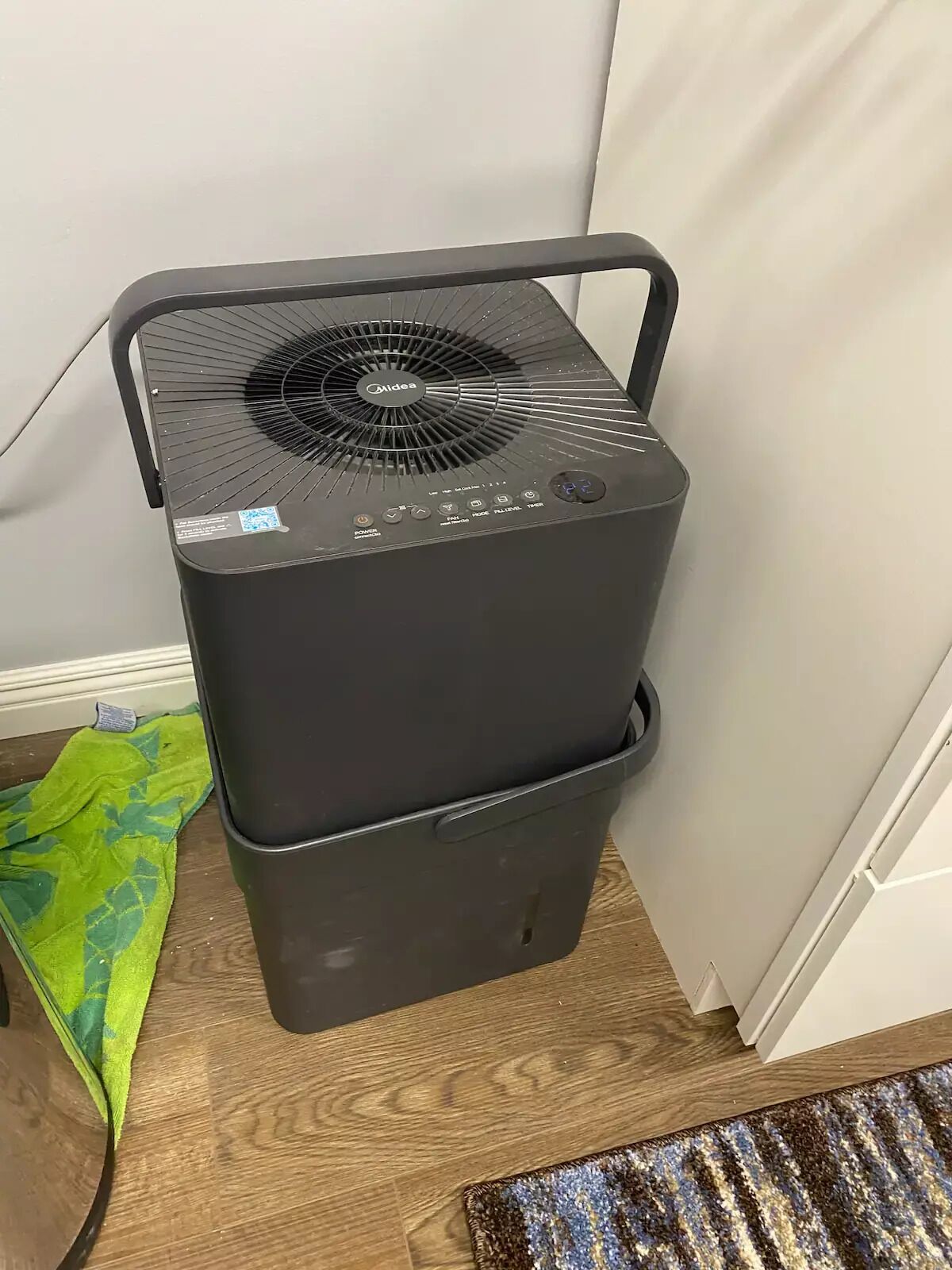Un termosifone portatile nero con un pannello di controllo e un cavo di alimentazione, posizionato su un pavimento in legno accanto a un asciugamano verde e un tappeto decorato.