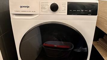 Une machine à laver blanche, fixée au mur, dotée d’un panneau de commande et d’un couvercle rouge.