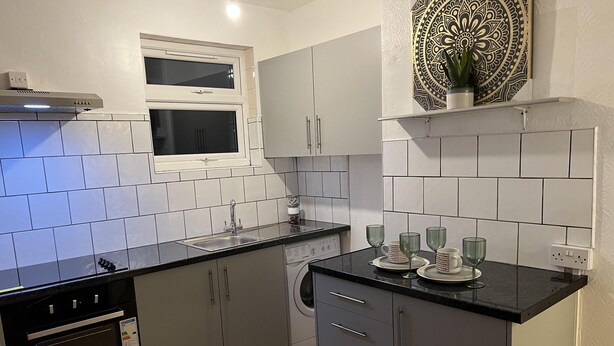 A compact kitchen with a black countertop, stainless steel sink, and a built-in oven. There are grey cabinets and a decorative wall piece above the counter. - Apartment in Leicester- Doublebed-citycentre-wifi (Leicester)