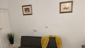 A living room with a black sofa, a yellow throw blanket, a small yellow side table with a plant, and two framed pictures on the wall. - Apartment in Leicester- Doublebed-citycentre-wifi (Leicester)