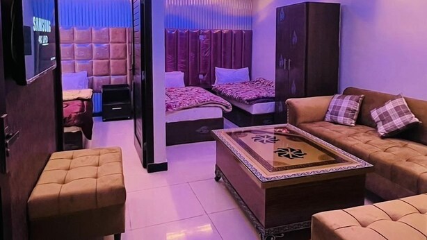 A modern living room with a sofa, ottoman, and a coffee table. There is a TV mounted on the wall, and a ceiling fan is visible. - Hotel 11 (Islamabad)